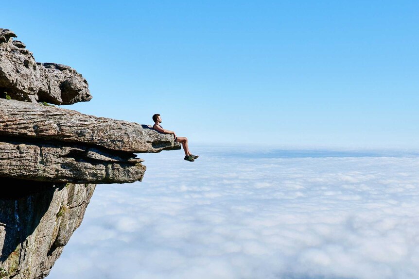 Man sits on the edge of a cliff rock for a story about why we take risks when travelling overseas.