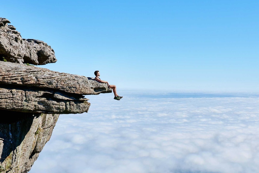Man sits on the edge of a cliff rock for a story about why we take risks when travelling overseas.
