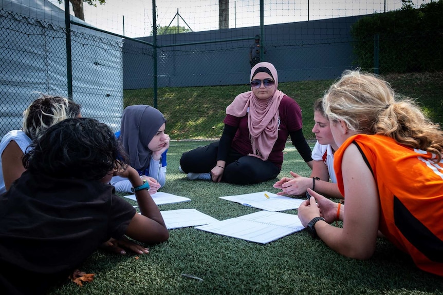 A woman sits on the grass with some junior football players, discussing strategy for the game.