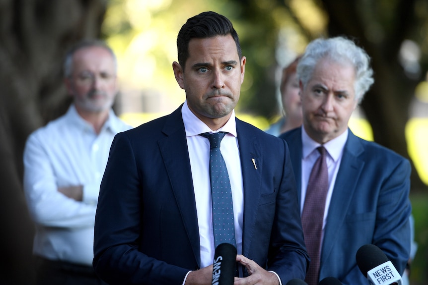 A man looking glum stands in front of several microphones. 