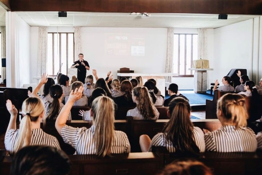 A room of students raise their hands as a man addresses them