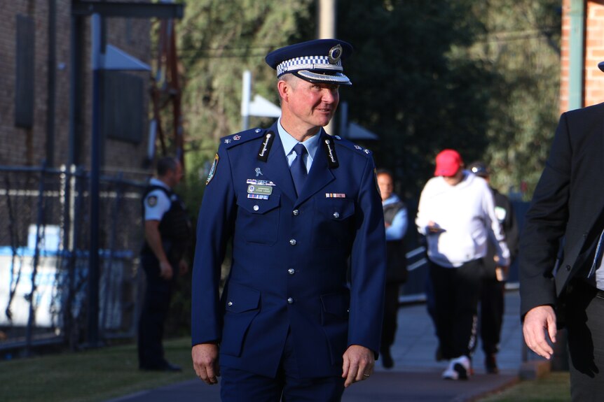 Man in formal navy police suit and cap, looks away from the camera as he walks towards it