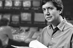 Black and white photo of Olle holding script with television monitors in background.