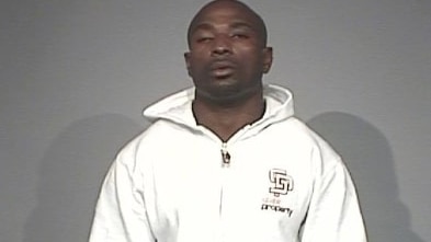 A Nigerian man stands for a mug shot wearing a full white tracksuit. He is visible from the waist up.