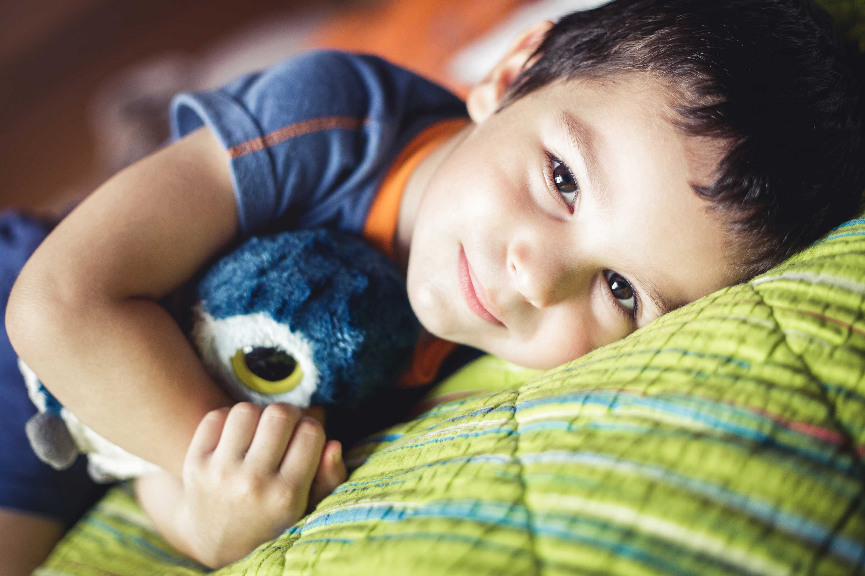 Young boy lying on his side looking into the camera lens. He is cuddling a soft toy owl. 