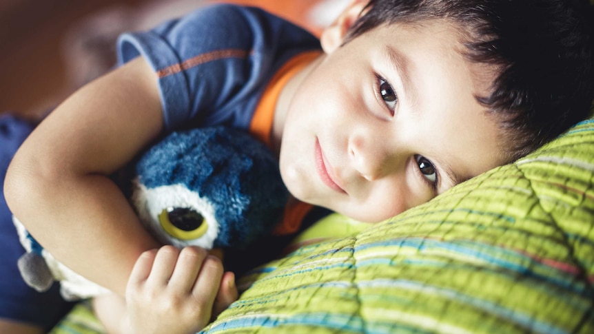 Young boy lying on his side looking into the camera lens. He is cuddling a soft toy owl. 