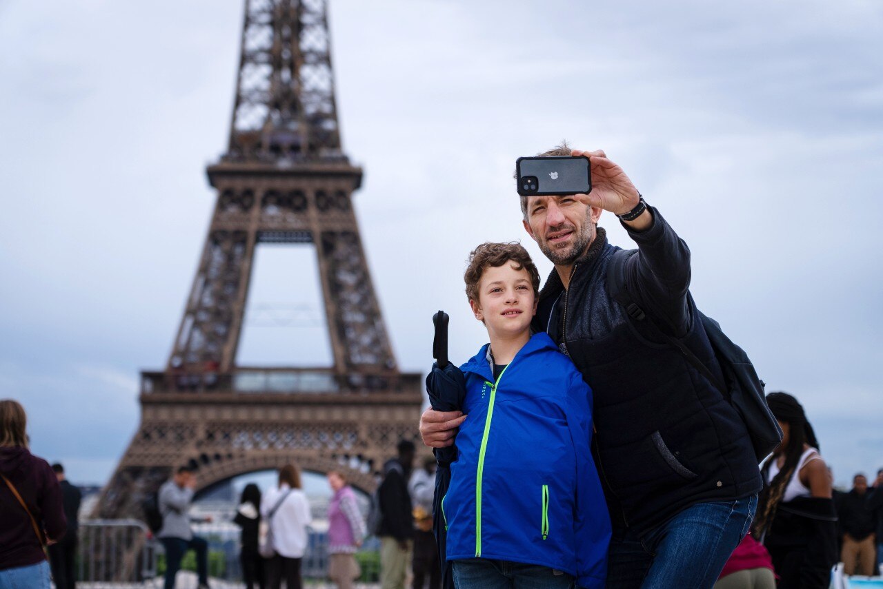A man and child stand in front of the Eiffel Tower in Paris, taking a selfie.