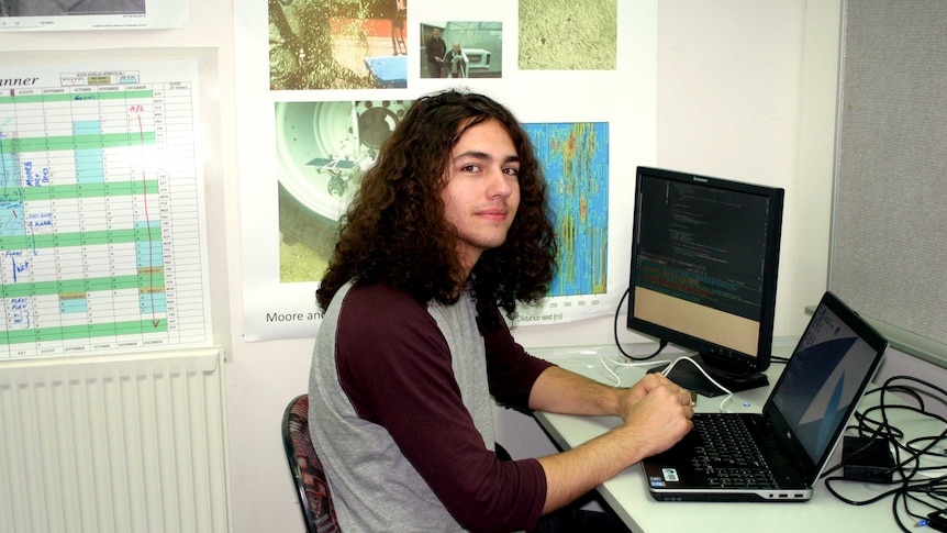 A young man sits at a desk with two computer screens
