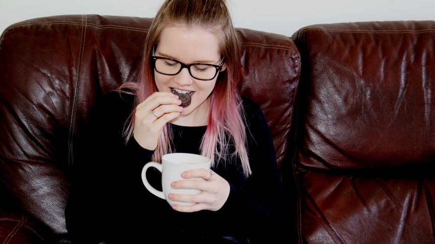 A woman sits on a couch eating an Oreo biscuit with a cup of tea.