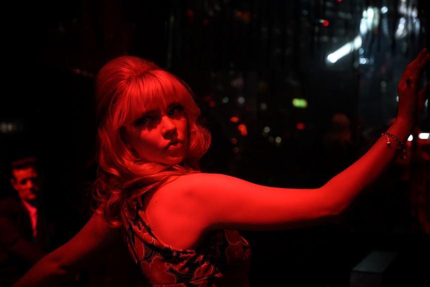 Bathed in a red light, a blonde woman in 60s clothes dances