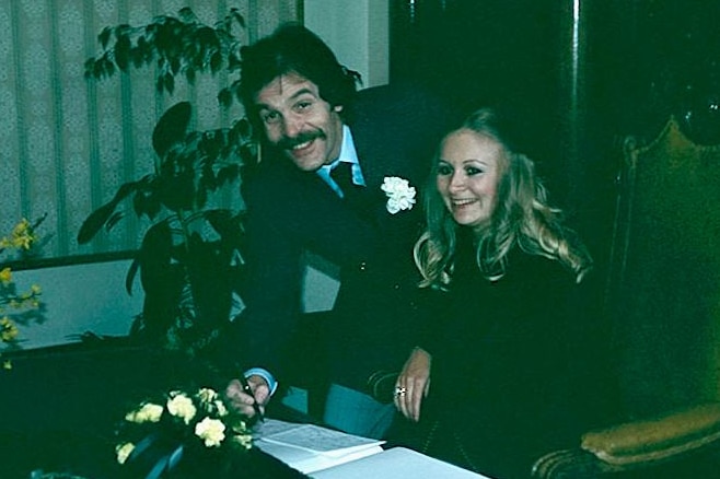 A couple on their wedding day sign documents, with a suited man with a moustache and a blonde woman.