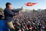 A man waves the flag of Kyrgyzstan as he looks over a large crowd of protesters who face him.