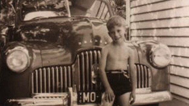 A young barefoot boy in a pair of shorts leans against the front of an old car parked next to a house.