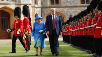 Queen dressed in blue walks next to Trump on green lawn past a line of guards in red jackets holding bayonets.