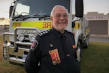 A senior man with white hair smiles with pride at the camera. He wears service medals and stands in front of a fire truck.