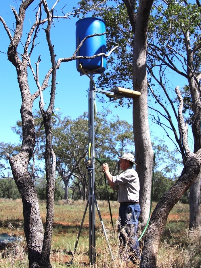 A man installs a water station on a tall pole in the bush.