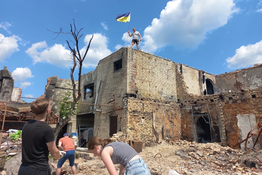 A man stands with a Ukrainian flag at the top of a damaged brick building