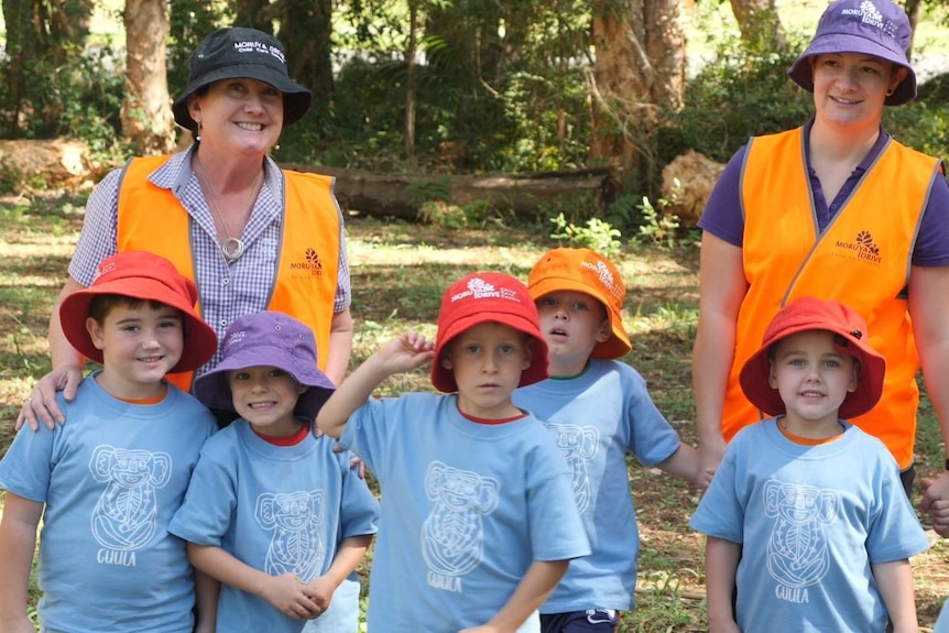 A group of preschool students in colourful hats and two adults wearing hats and hi-viz vests stand in the bush, smiling.
