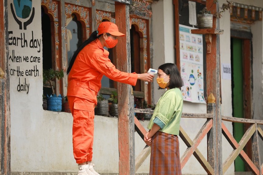 A woman wearing orange trousers and tops, an orange mask and a hat has a thermometer on her head.