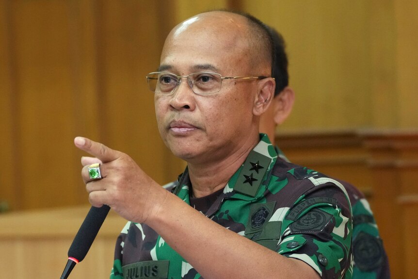A bald man in a military camo wearing a green ring points. 
