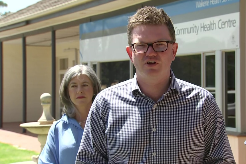 A man wearing a checked shirt in front of a building with a woman and a man behind him