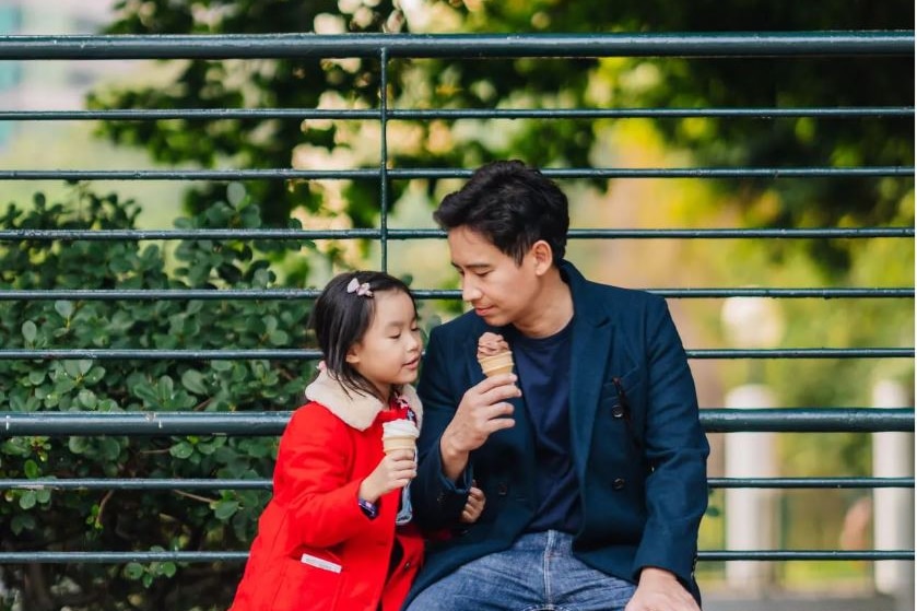 A man wearing  a dark blue coat holds a chocolate icecream as he looks down at his daughter in a red coast holding an icecream.