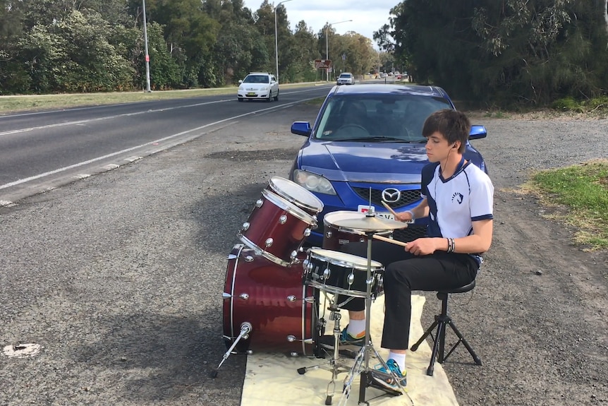 An 18-year-old boy sits at a drum kit on the side of the road, playing as cars pass by.