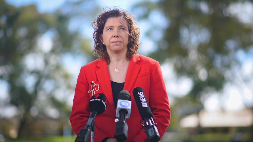A woman with curly brown hair wearing a red suit jacket stands outside in front of an array of microphones.