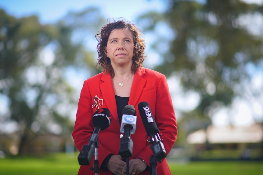 A woman with curly brown hair wearing a red suit jacket stands outside in front of an array of microphones.