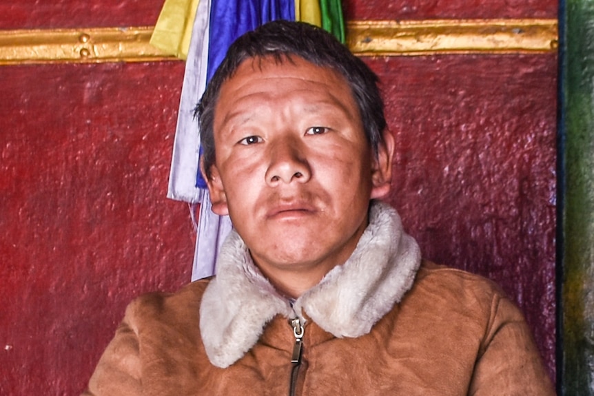 A man looks sad in a sheepskin jacket outside a colourful monastery