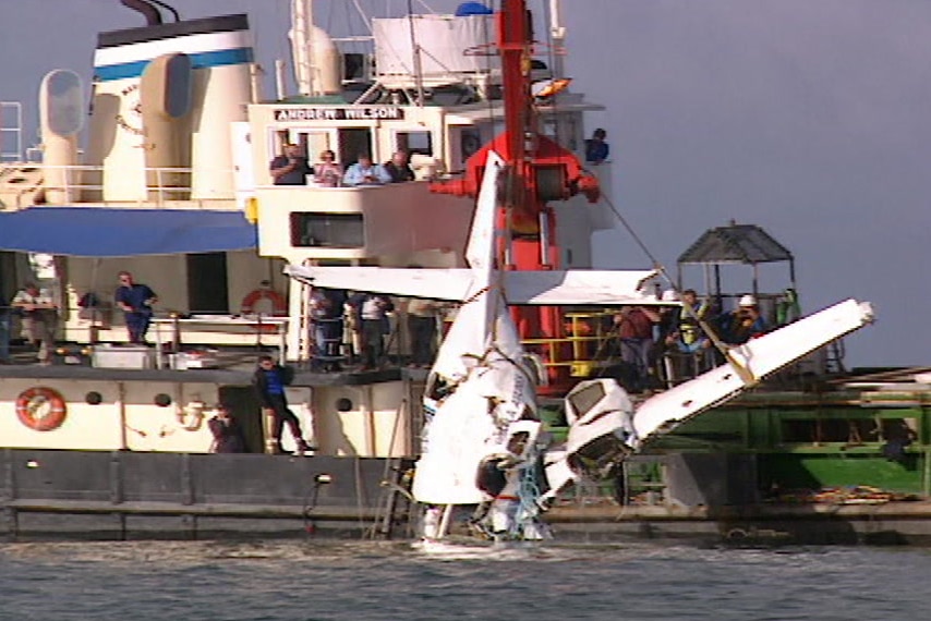 The wreckage of a plane is pulled out of the water and onto a boat. The top of the plane is missing.