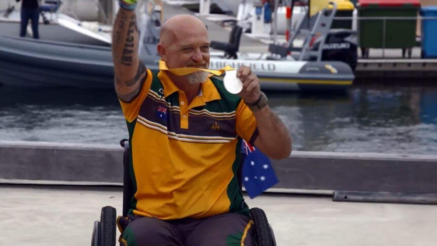 A man sits in a wheelchair with an Australian flag, holds up a silver medal and puts his tattooed arm in the air in celebration.