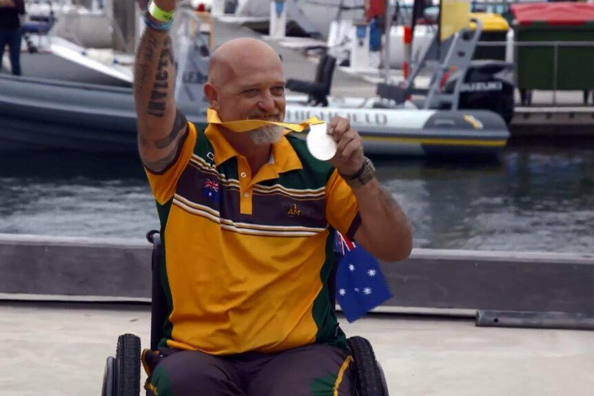 A man sits in a wheelchair with an Australian flag, holds up a silver medal and puts his tattooed arm in the air in celebration.