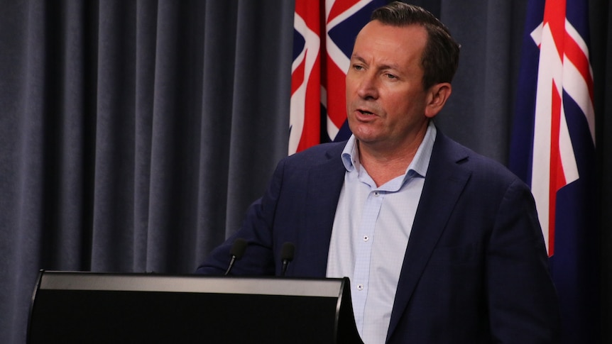 A side-shot of Mark McGowan standing at a podium in front of a blue curtain and Australian flag, wearing a suit.