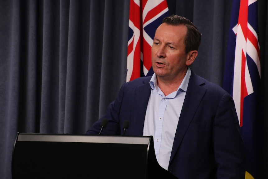 A side-shot of Mark McGowan standing at a podium in front of a blue curtain and Australian flag, wearing a suit.