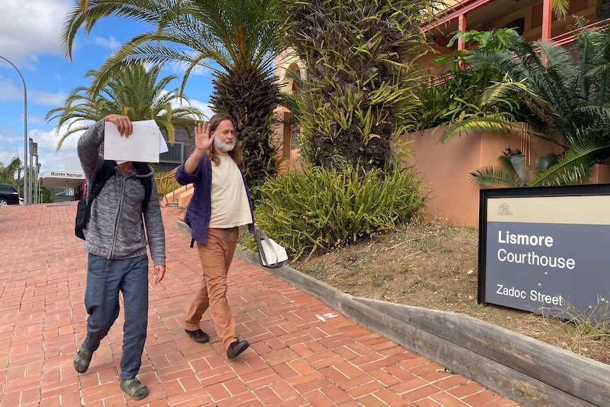 Two men hold their hands up to the camera as they walk past a sign that says "Lismore Courthouse".