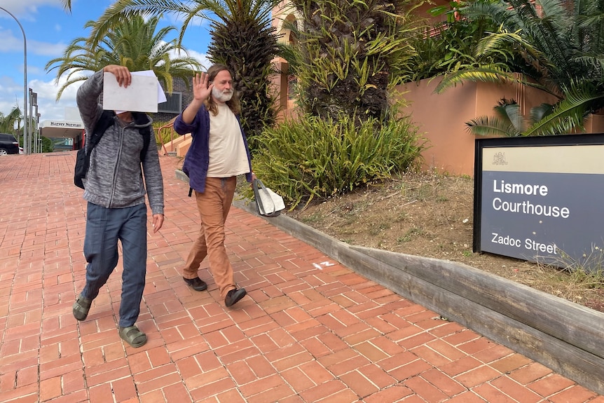 Two men walk past Lismore Courthouse sign - one with a notepad held over his face.