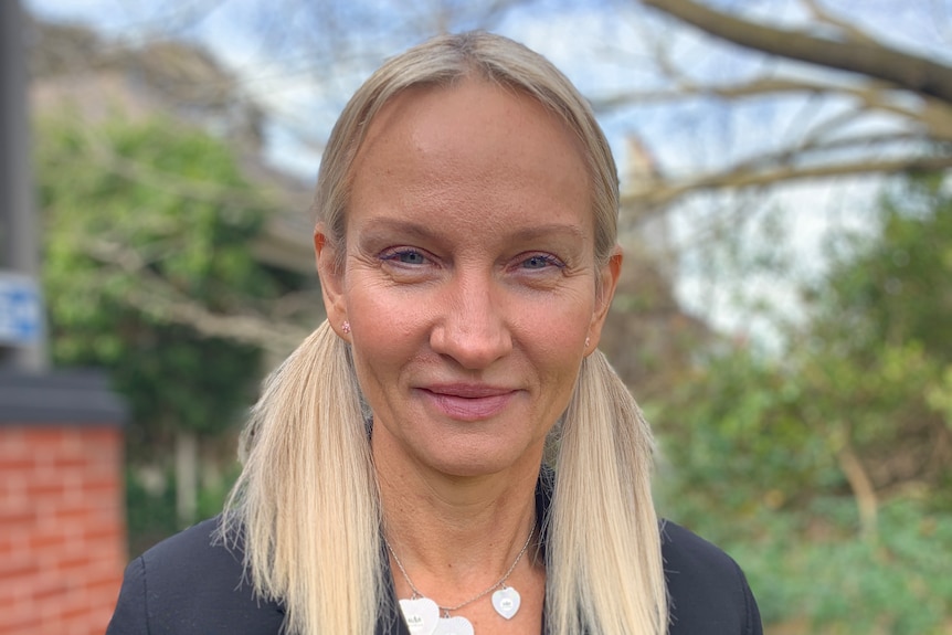 A woman with blonde hair stands outside with a tree behind her. She is smiling. 