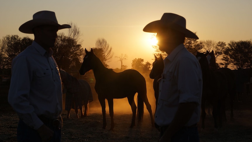 Two men wearing cowboy hats stand near horses at sunrise.
