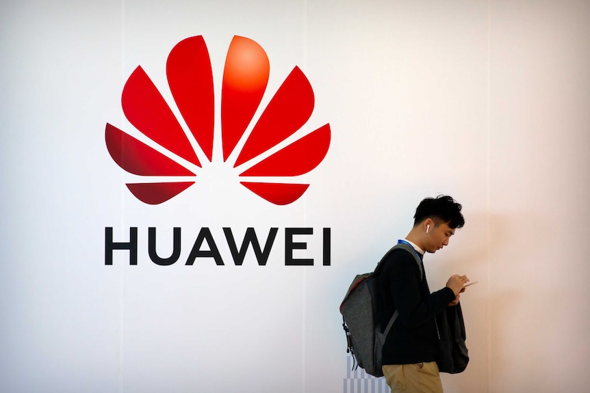 A man on looking down at his phone walks past a Huawei logo