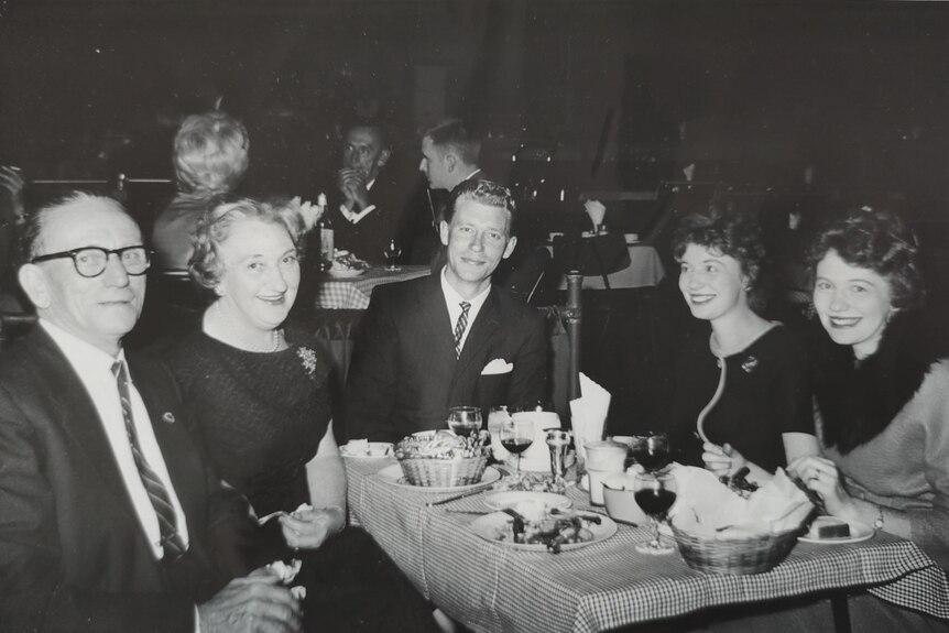 Old family photo of the twins at a dinner table with their parents and a young man in the middle.