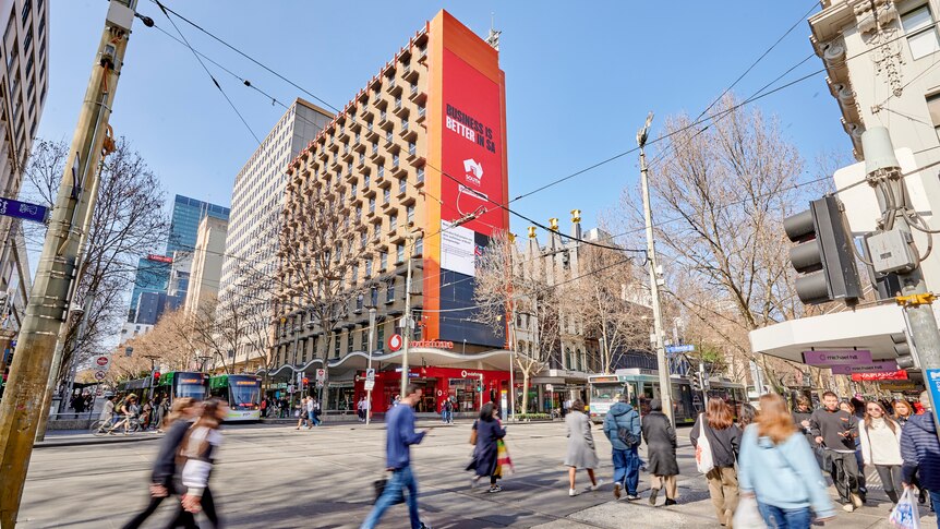 A large red billboard on a building on a busy street corner, with people walking past