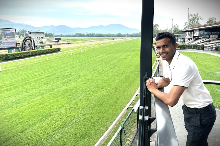 A young man with brown skin and black hair wearing a white shirt and black pants overlooks a lush, green race track.