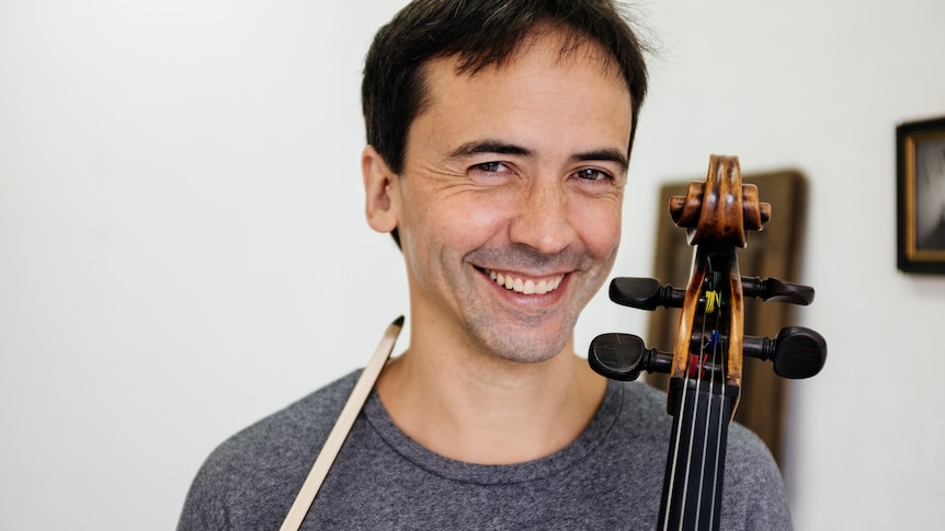 A man smiles at the camera. A cello scroll and bow rest against his shoulders.