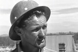 A black and white still of a man wearing a hard hat speaking to Four Corners