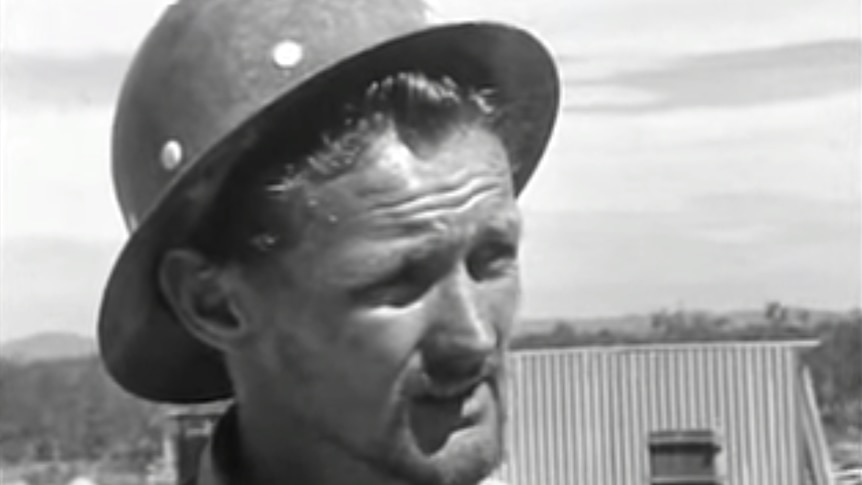 A black and white still of a man wearing a hard hat speaking to Four Corners