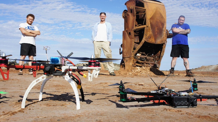 Three Goldfields drone enthusiasts pose with their flying craft in the foreground and a rusted car body.