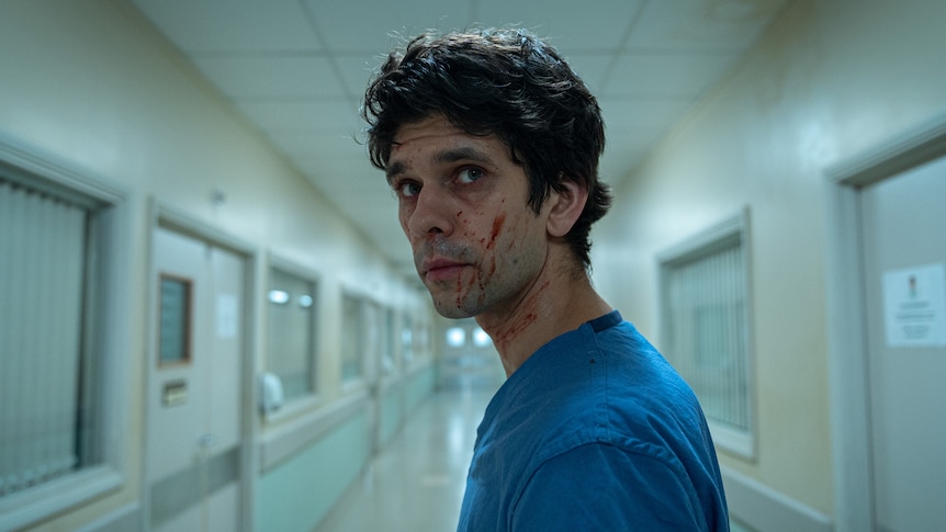 A TV still of Ben Whishaw. He's turning to look down a hospital corridor behind him, and has blood splattered on his face.