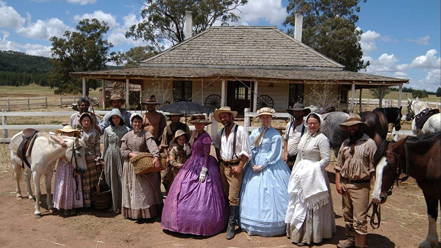 People in period dress stand in front of homestead
