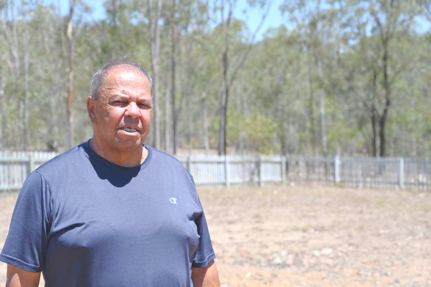 An aboriginal man wearing a blue shirt stands, serious with a dry brown grass covered field in the background.